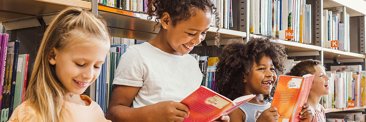 Group of four children, three girls and a boy smiling while standing in front of book stacks and holding open books