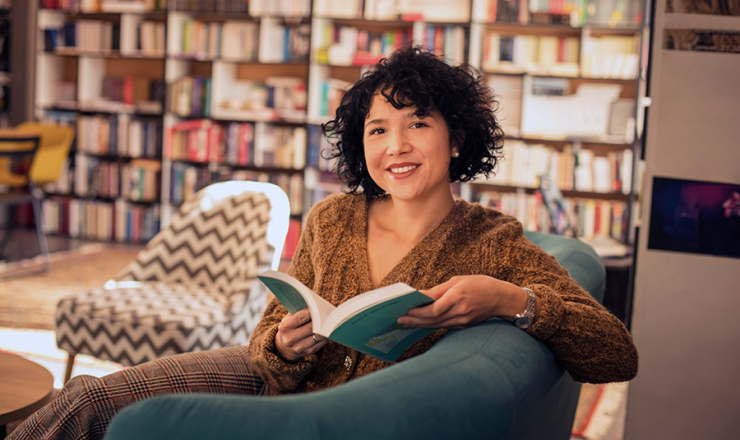 Smiling woman sitting on sofa in library with open book in her hands