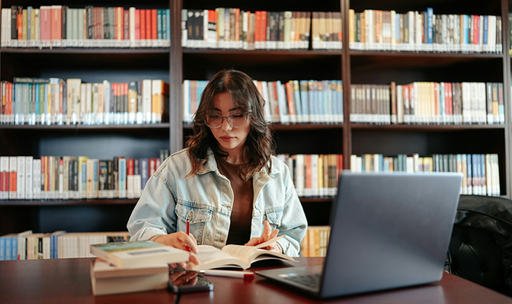 Teen girl with stack of books and open laptop on desk inside what looks to be a library